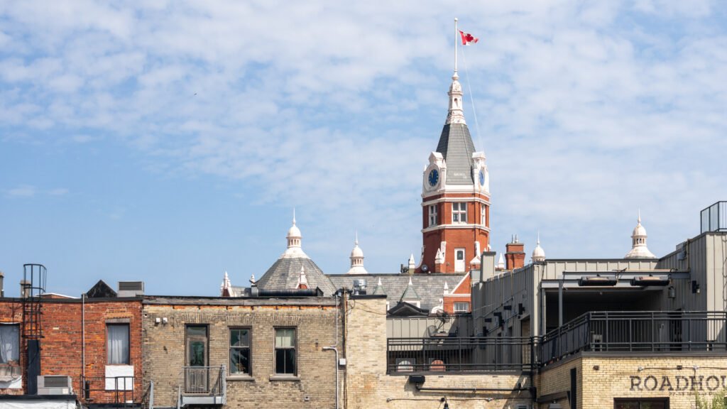 Red brick clock tower with a Canadian flag atop, rising above rooftop buildings against a blue sky.