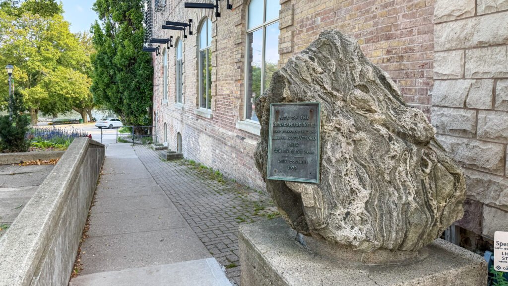 History Stratford | Krista the Explorer Rock with a bronze plaque beside a brick building along a paved walkway.