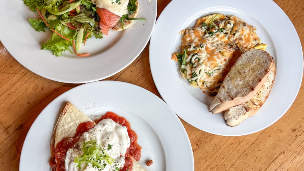 Foster’s Inn Stratford | Krista the Explorer Three plates of food on a wooden table: a salmon salad sandwich, a cheesy omelette with toast, and huevos rancheros.