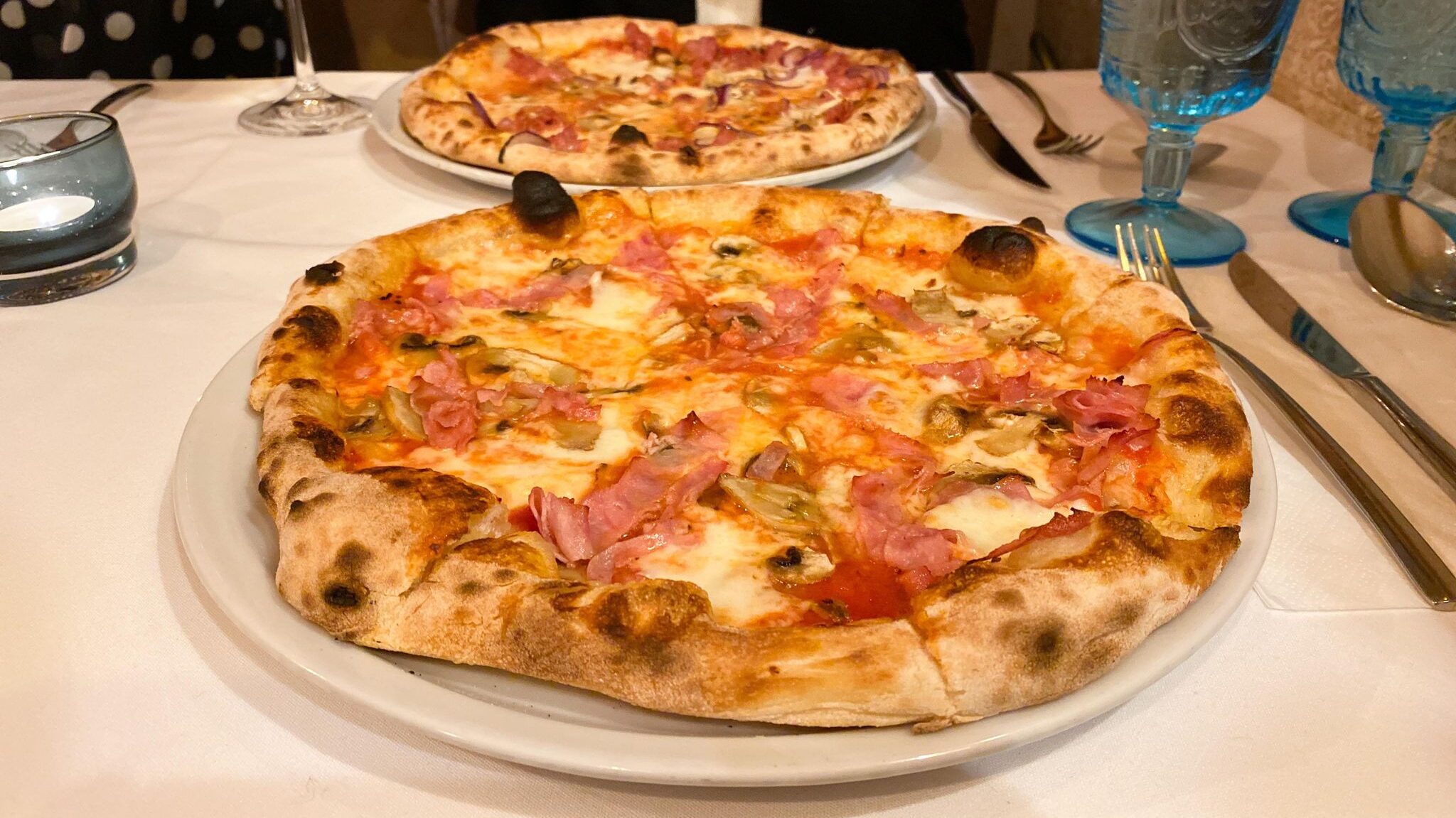 Two stone baked pizzas on white tablecloth. 