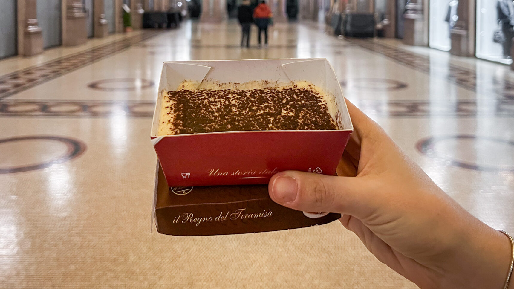 Classic tiramisu in cardboard container inside shopping arcade.