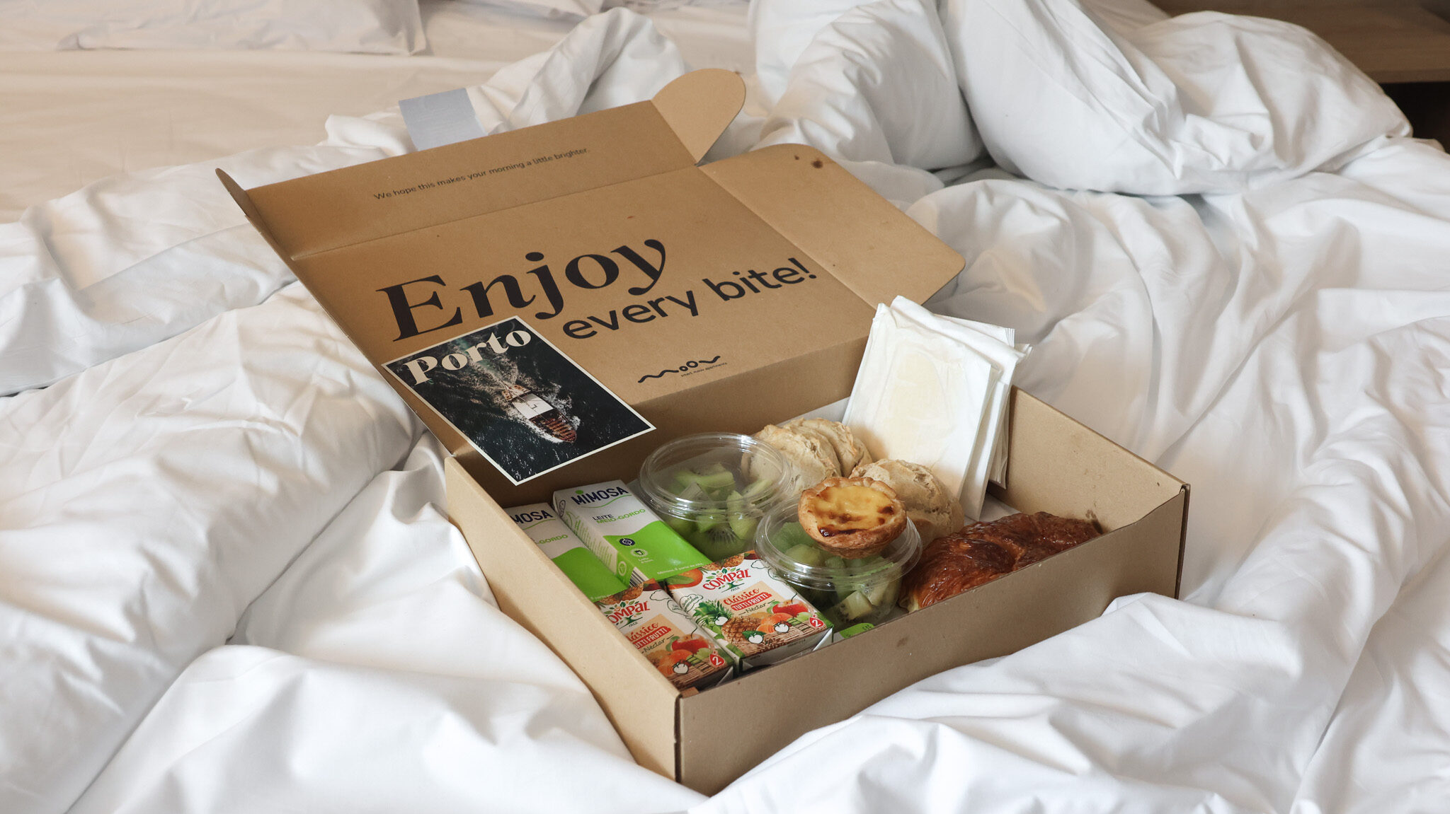 Box of breakfast items sitting on messy bedding.