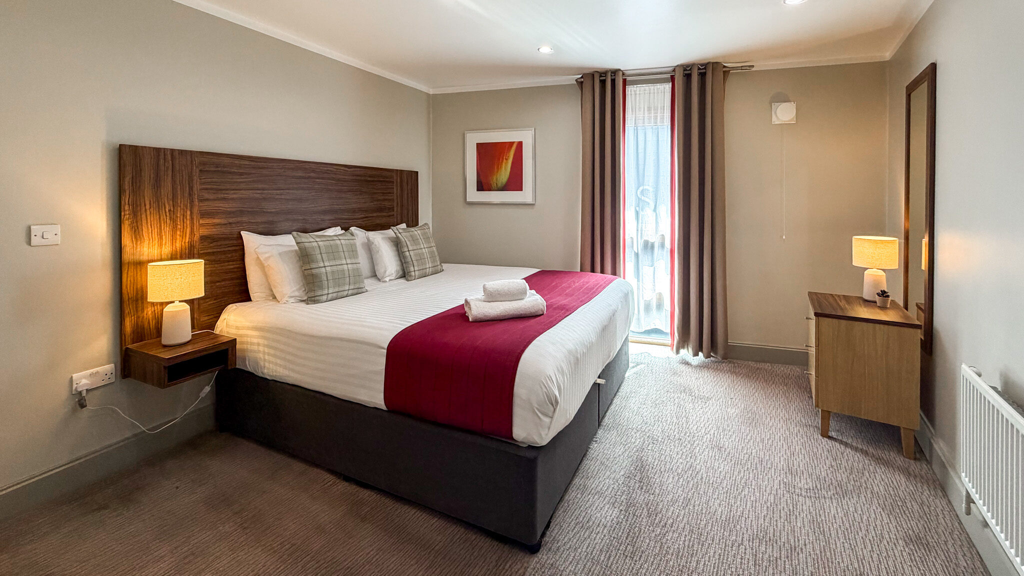 Hotel-style bedroom with a large wooden headboard bed, white linens, burgundy throw, and plaid pillows.