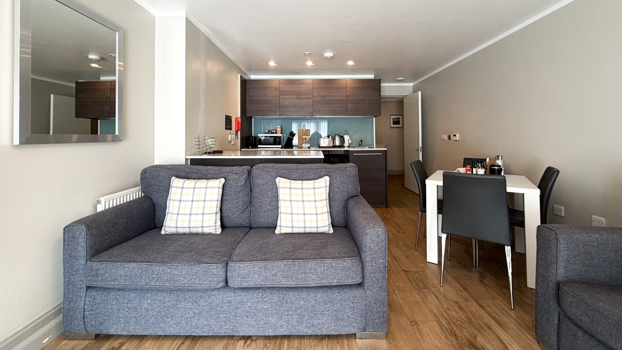 Living room with a gray sofa in the foreground, plaid cushions, and an open kitchen with dark wood cabinets in the background.