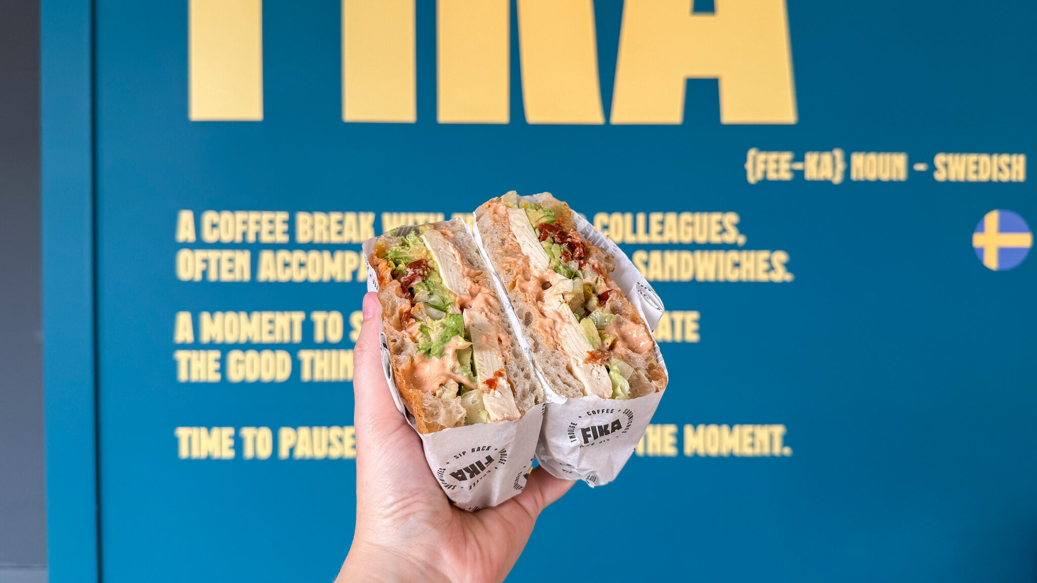 Hand holding a veggie-filled sandwich wrapped in paper against a blue wall.