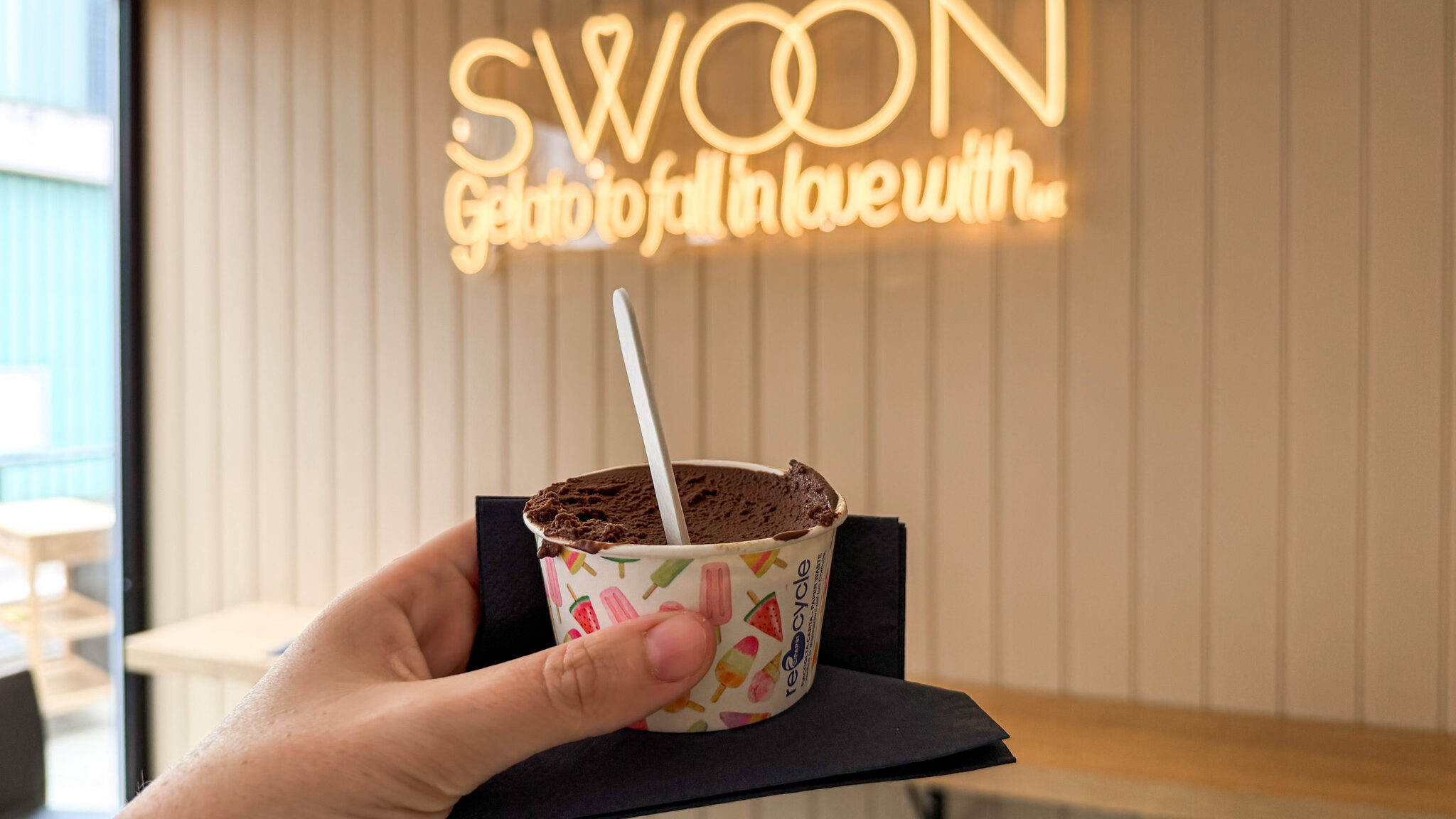 Hand holding a cup of chocolate gelato with a plastic spoon.