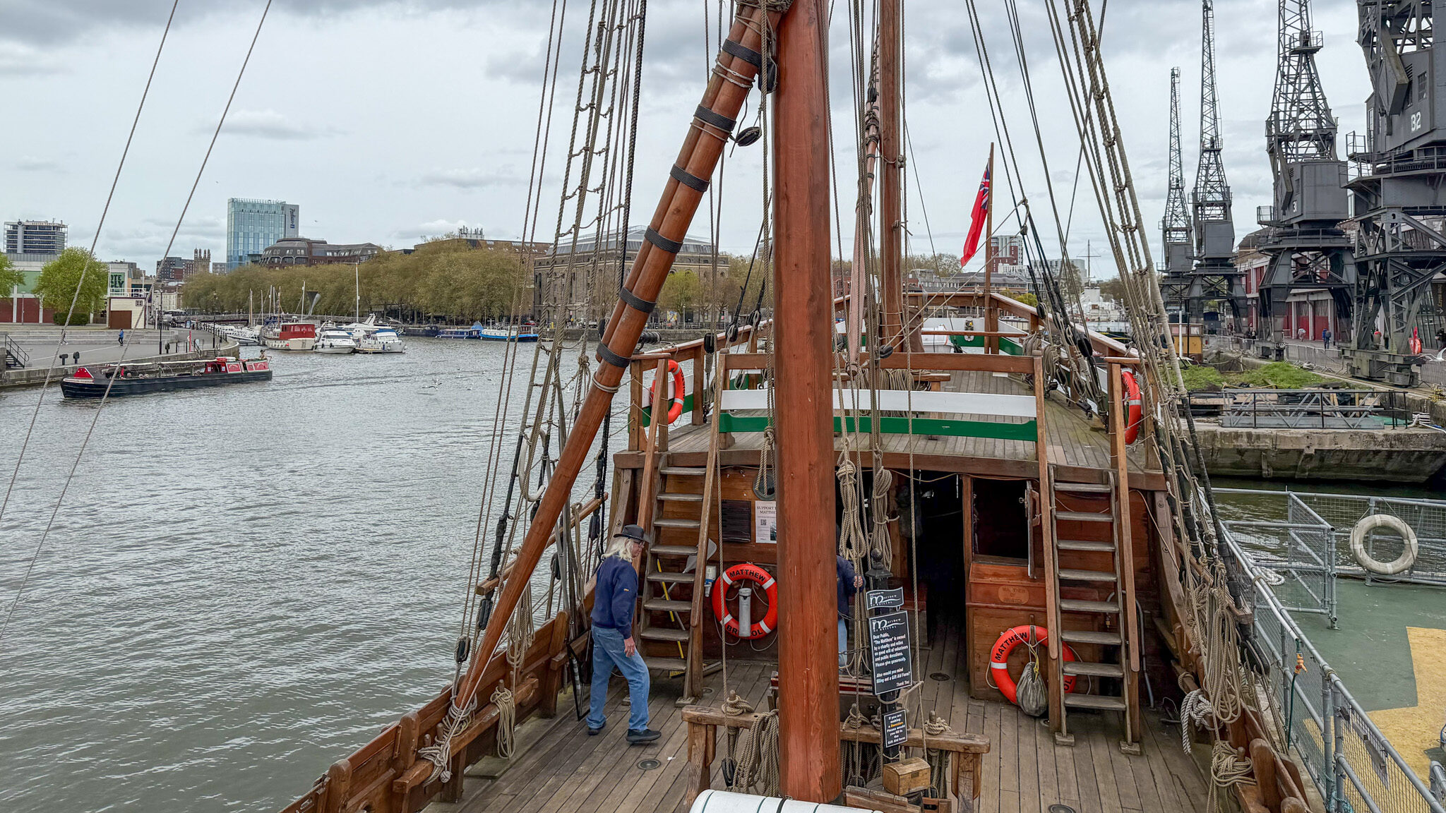 A wooden ship deck with tall masts and rigging, dockside warehouses and boats in the harbor beyond under cloudy skies.