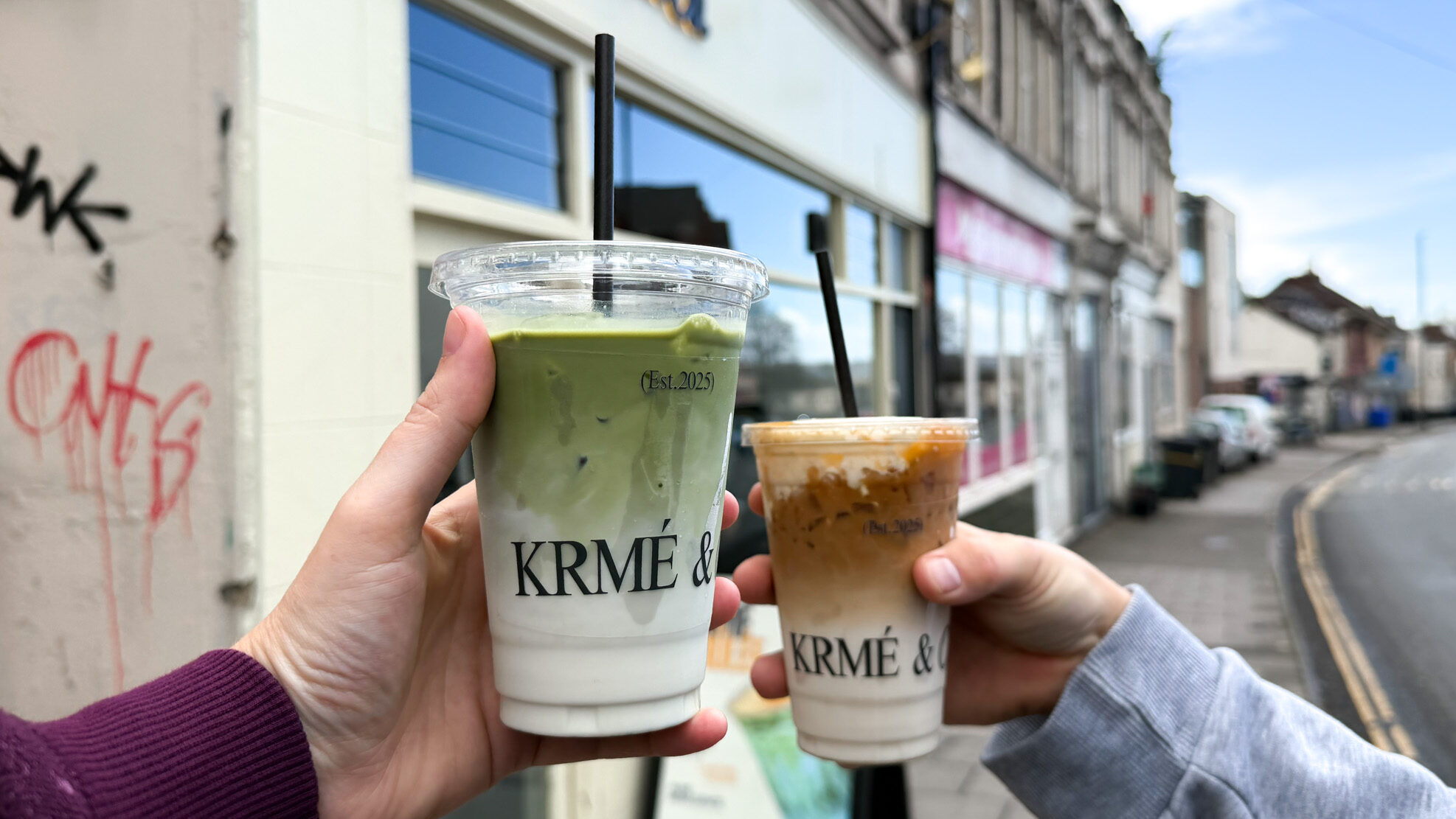 Matcha latte and iced coffee being held up in front of shop.