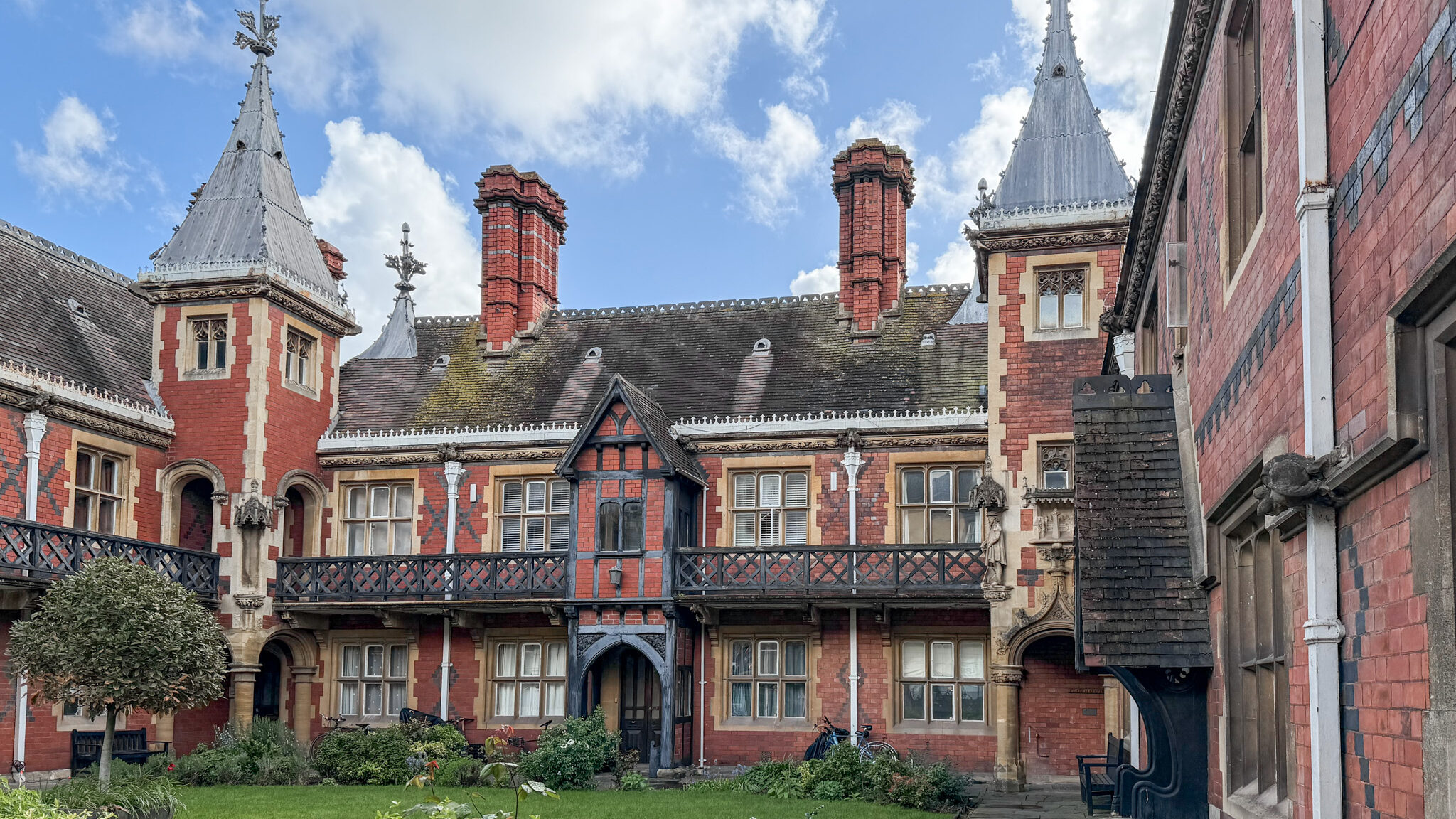 Red-brick historic building with steep slate roofs, decorative spires, and a small garden in the foreground.