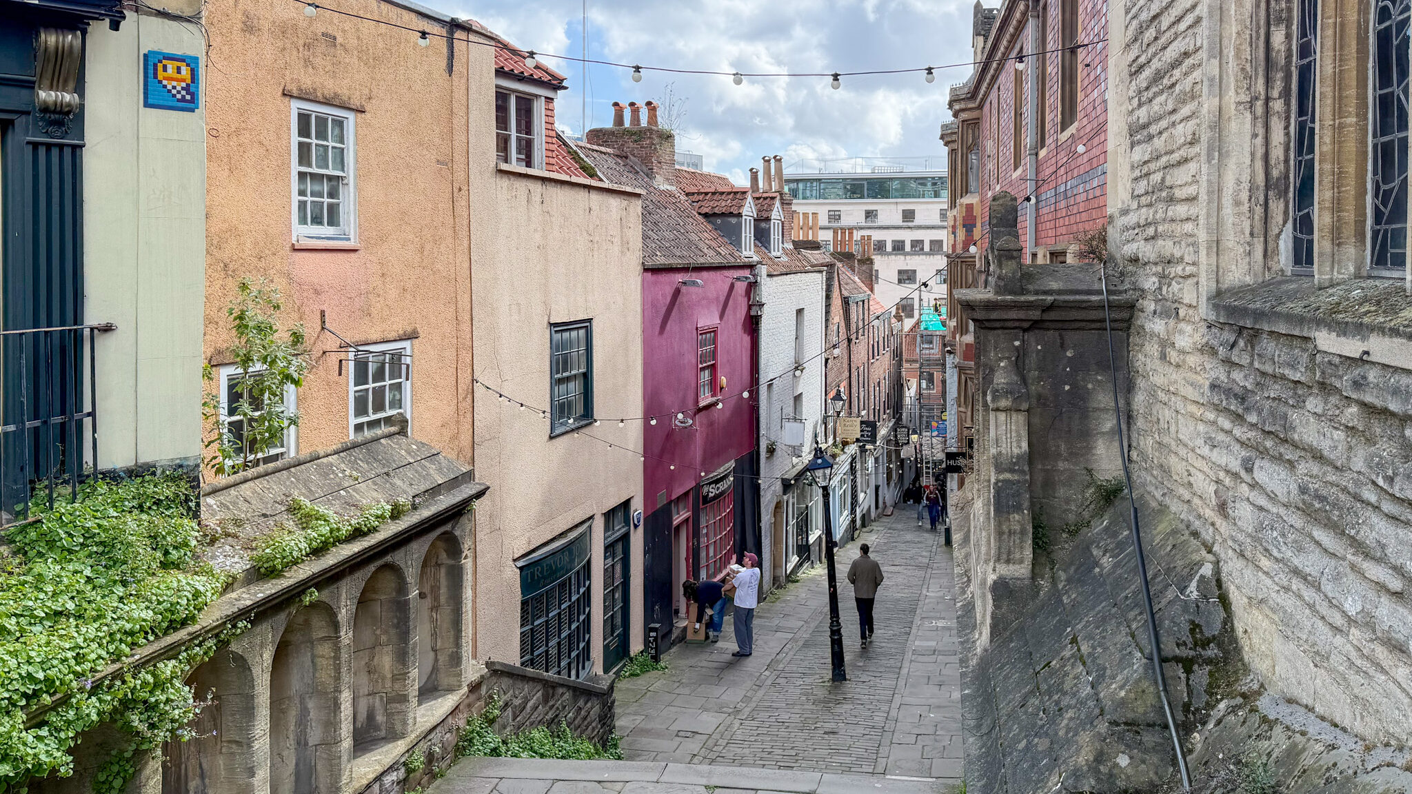 Narrow cobblestone street climbing uphill between pastel-colored houses with string lights overhead.