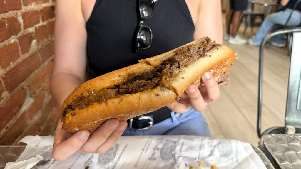 Person in a black shirt holds a long cheesesteak sandwich with beef and onions.