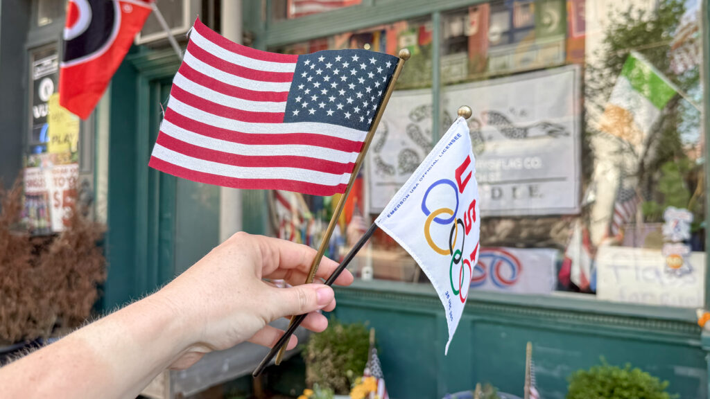 Close-up of a hand holding two small flags: the United States flag and an Olympic flag.