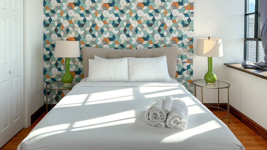 Bright bedroom with a geometric teal, orange, and white wallpaper behind a beige headboard.