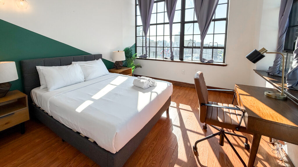 Bright bedroom with a large bed, white linens, and a wooden desk by a window with city views.