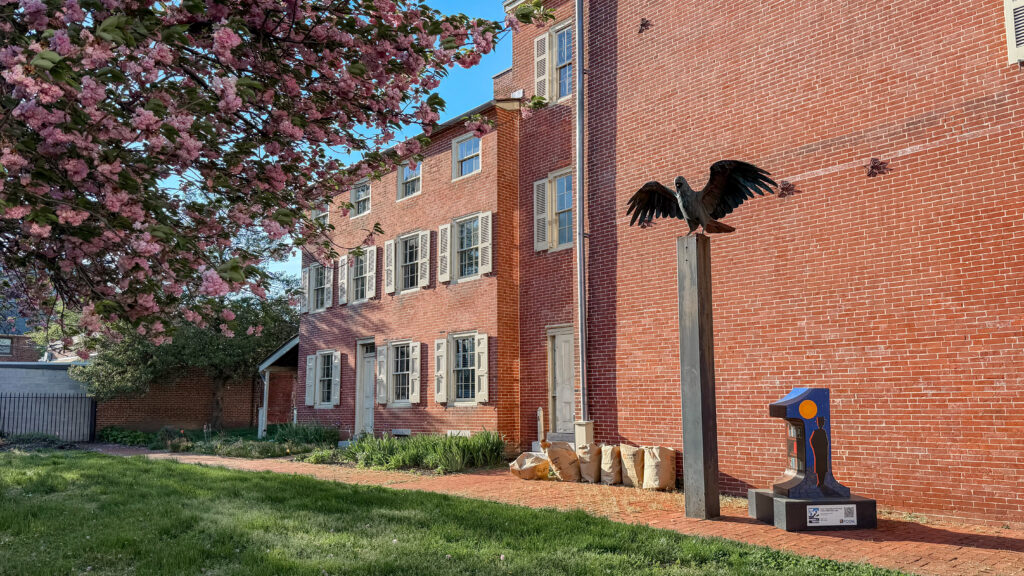 Red brick building with white-trimmed windows, pink flowering tree in the foreground, and a dark bird sculpture.