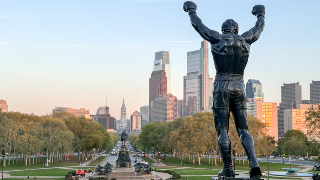 Bronze statue of a boxer with fists raised, overlooking a tree-lined park and Philadelphia skyline.