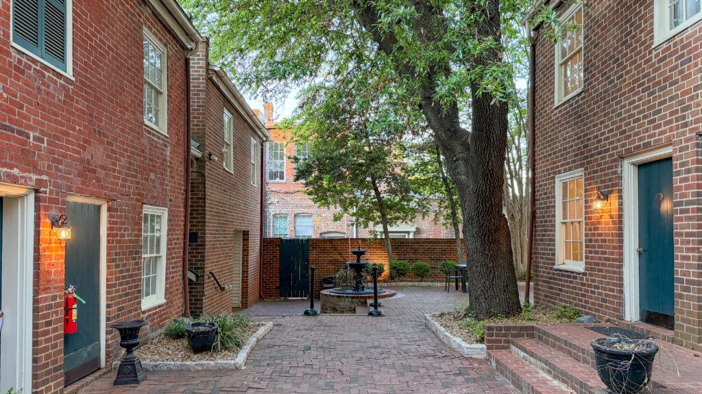 Brick courtyard between red brick buildings with a central fountain, trees, and outdoor seating edges.