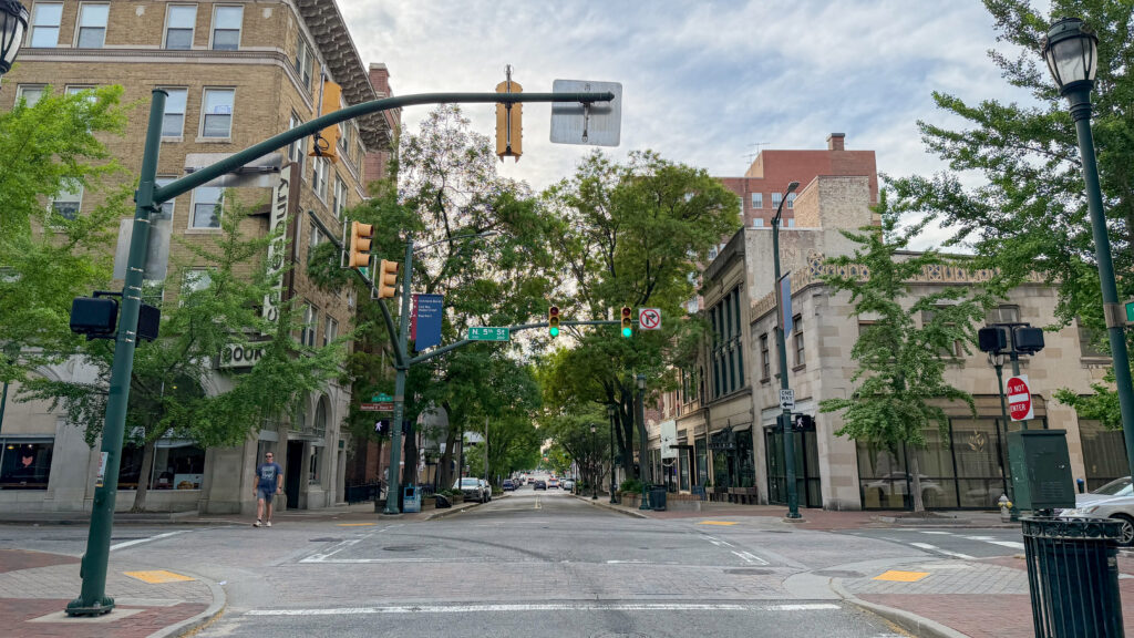 Urban street intersection with green traffic lights, brick and stone buildings, and trees lining the sidewalks.