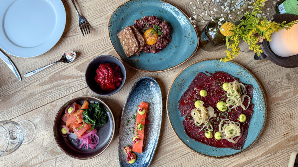 Top-down view of a wooden table set with blue ceramic plates featuring beef carpaccio with toppings and salmon.