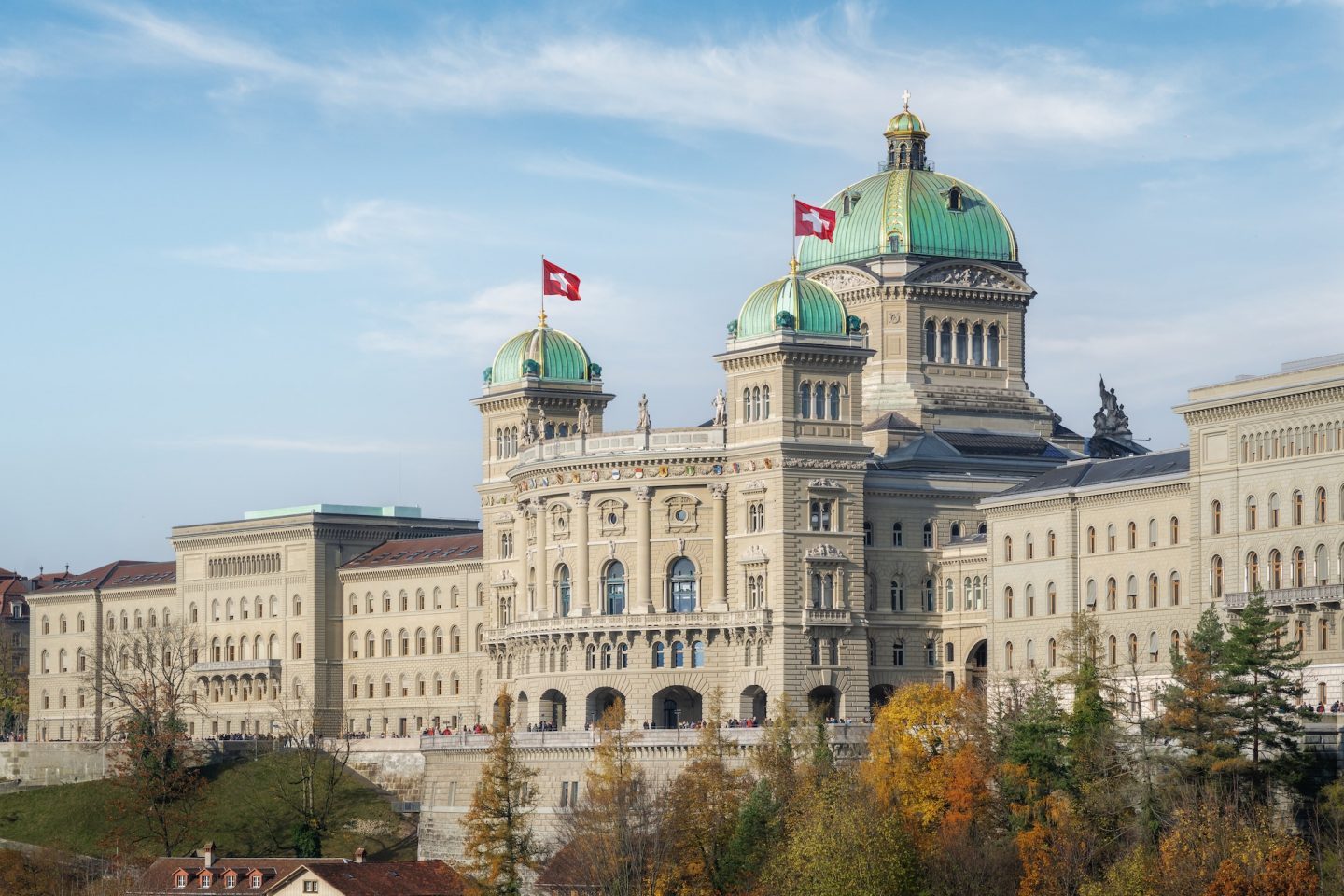 Grand government building with twin green domes topped by Swiss flags, set against a blue sky.