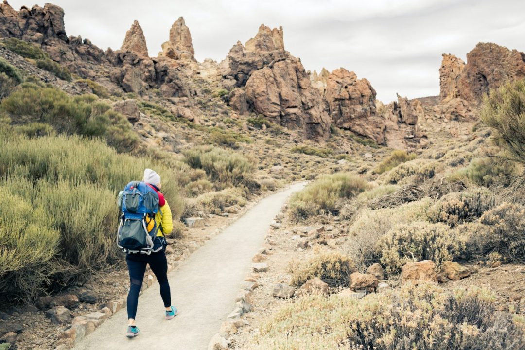 Hiker with a blue backpack walking a narrow desert trail toward jagged rock formations.