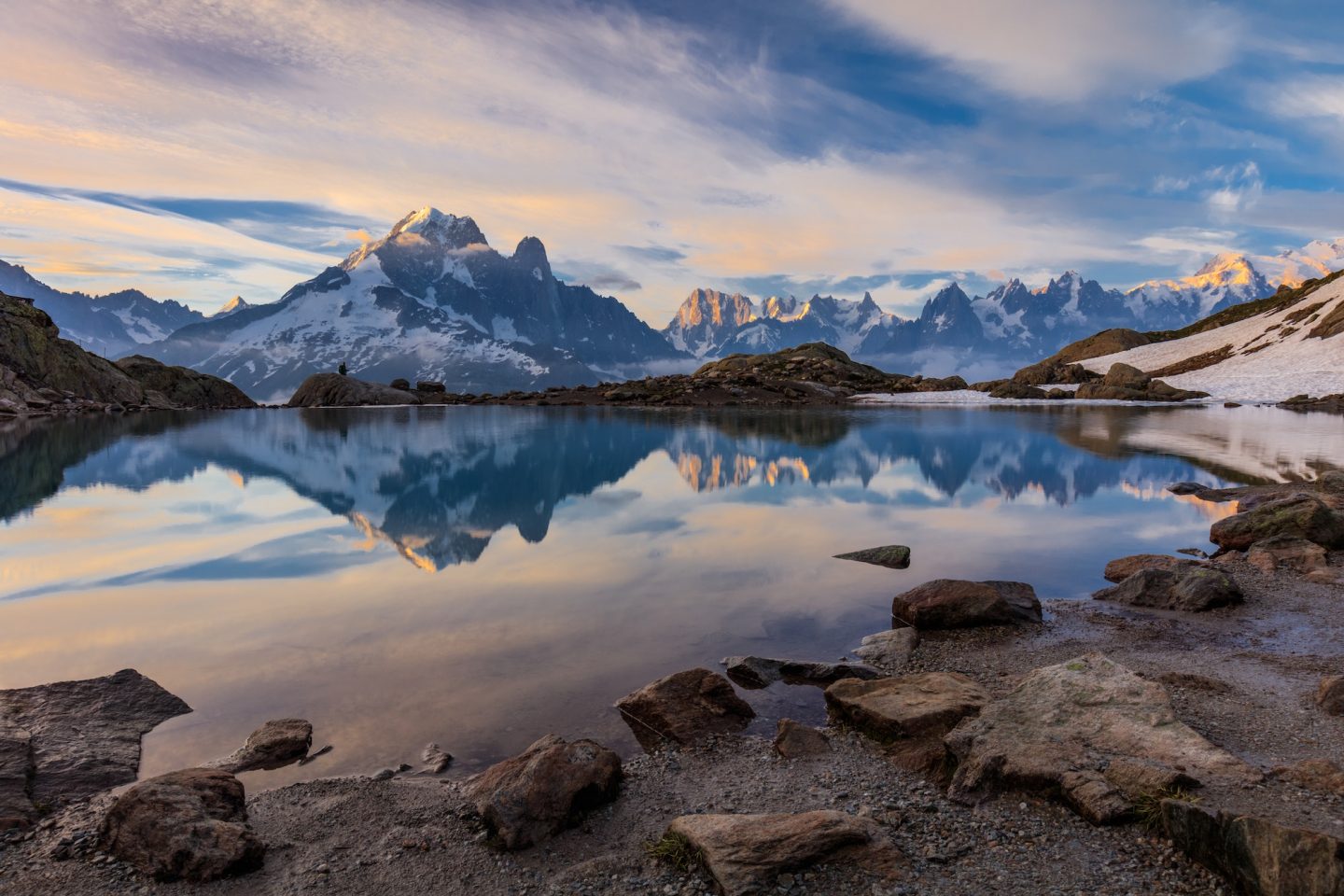 Alpine lake reflecting snow-capped mountains at sunset with a rocky shoreline in the foreground, calm and mirror-like water.