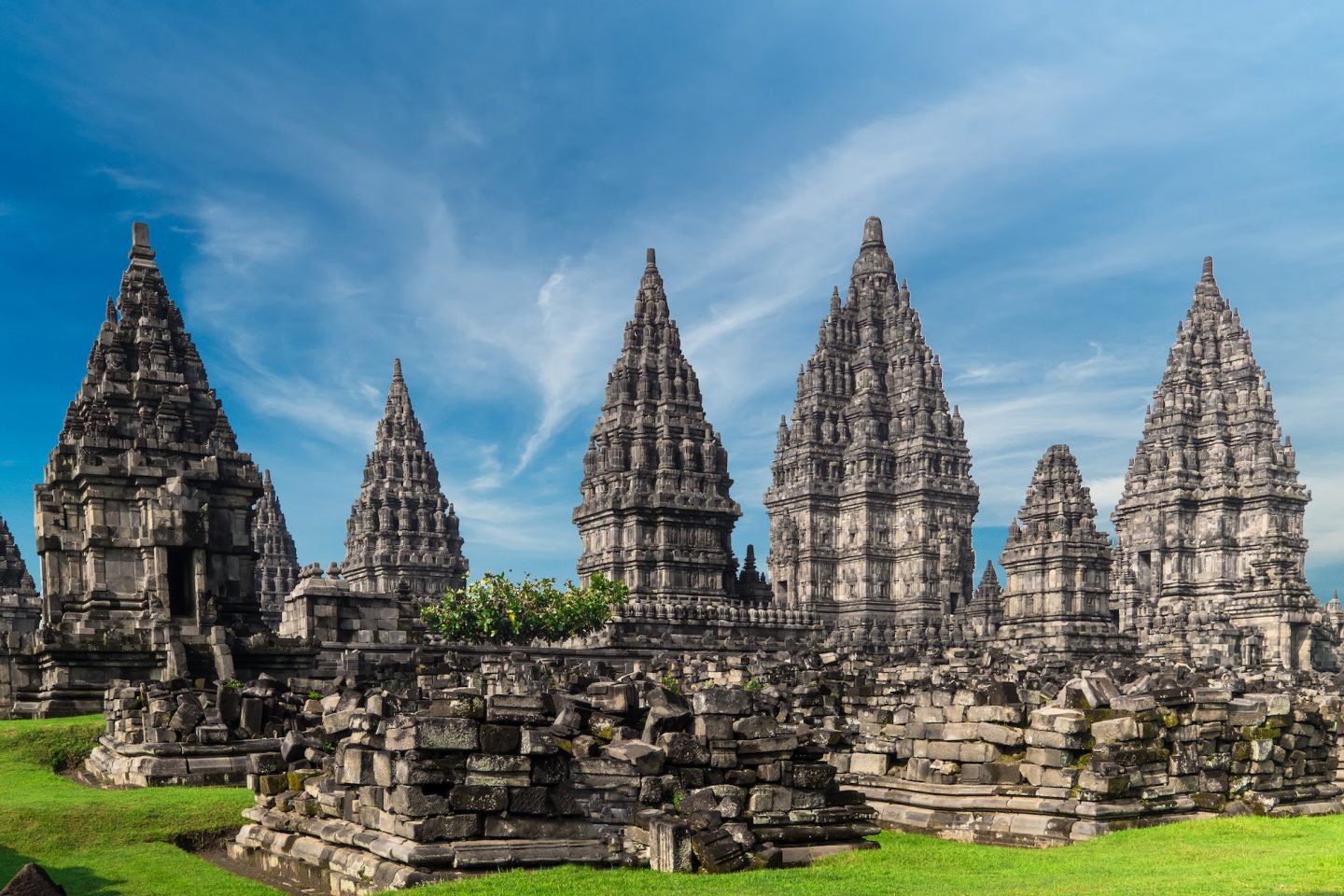 Ancient Hindu temple complex with tall, carved stone towers rising from a grassy courtyard under a blue sky.