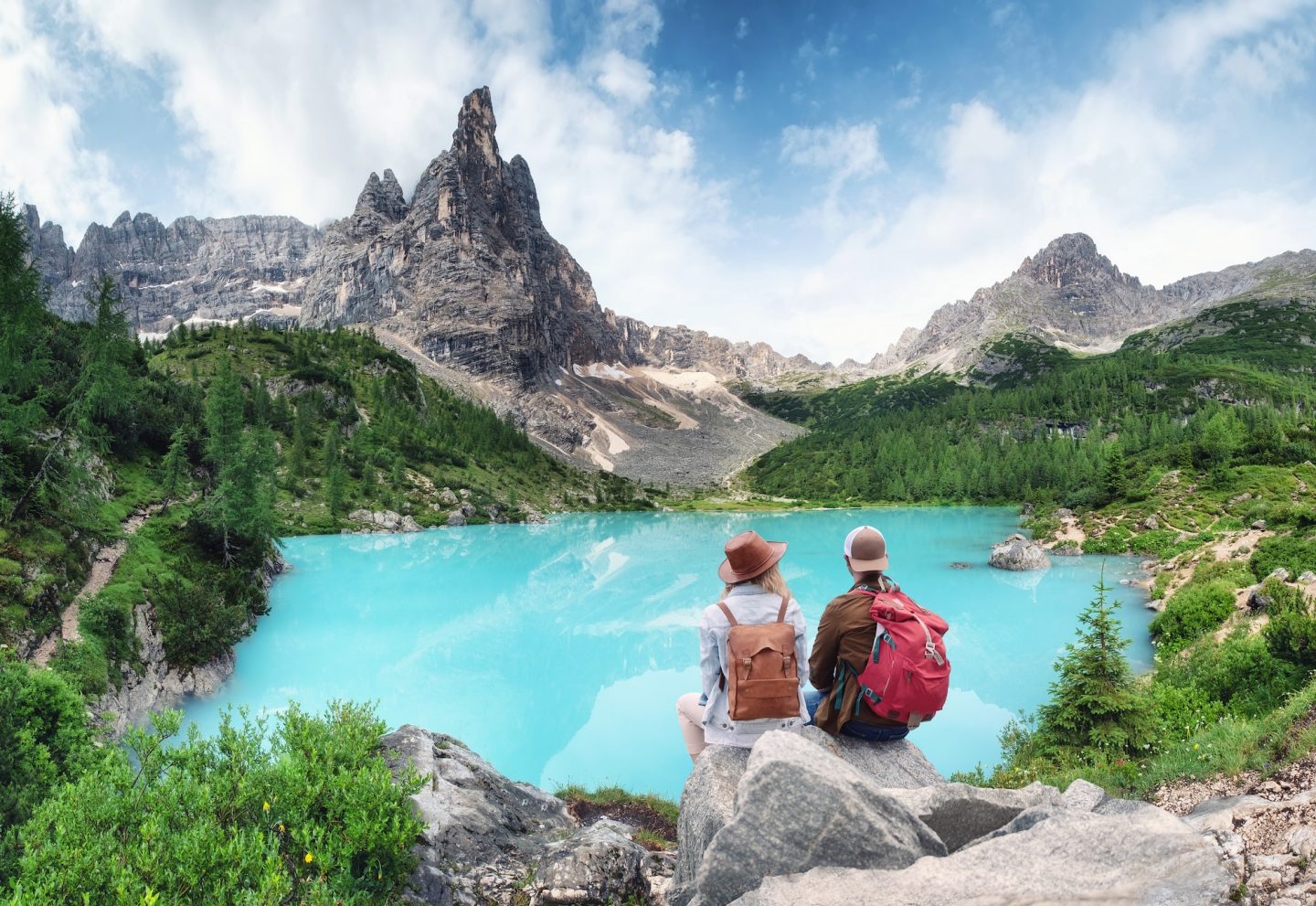 Two hikers with backpacks sit on a rocky ledge, overlooking a bright turquoise alpine lake and jagged mountain peaks.