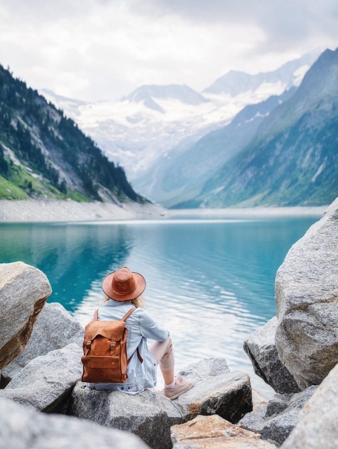Traveler with a brown backpack sits on rocks by a turquoise alpine lake, mountains in the distance.