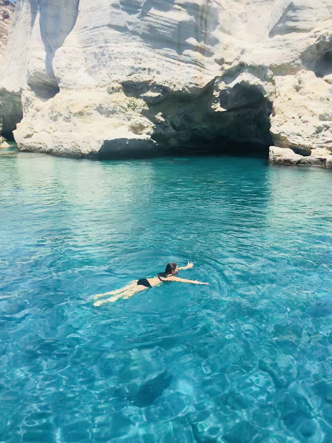 Person swimming in bright turquoise water near white rock cliffs with a sea cave in the background.