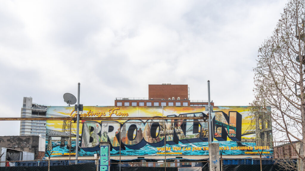 Street Art Brooklyn | Krista the Explorer Wide mural on a brick building reading'Greetings From Brooklyn'.