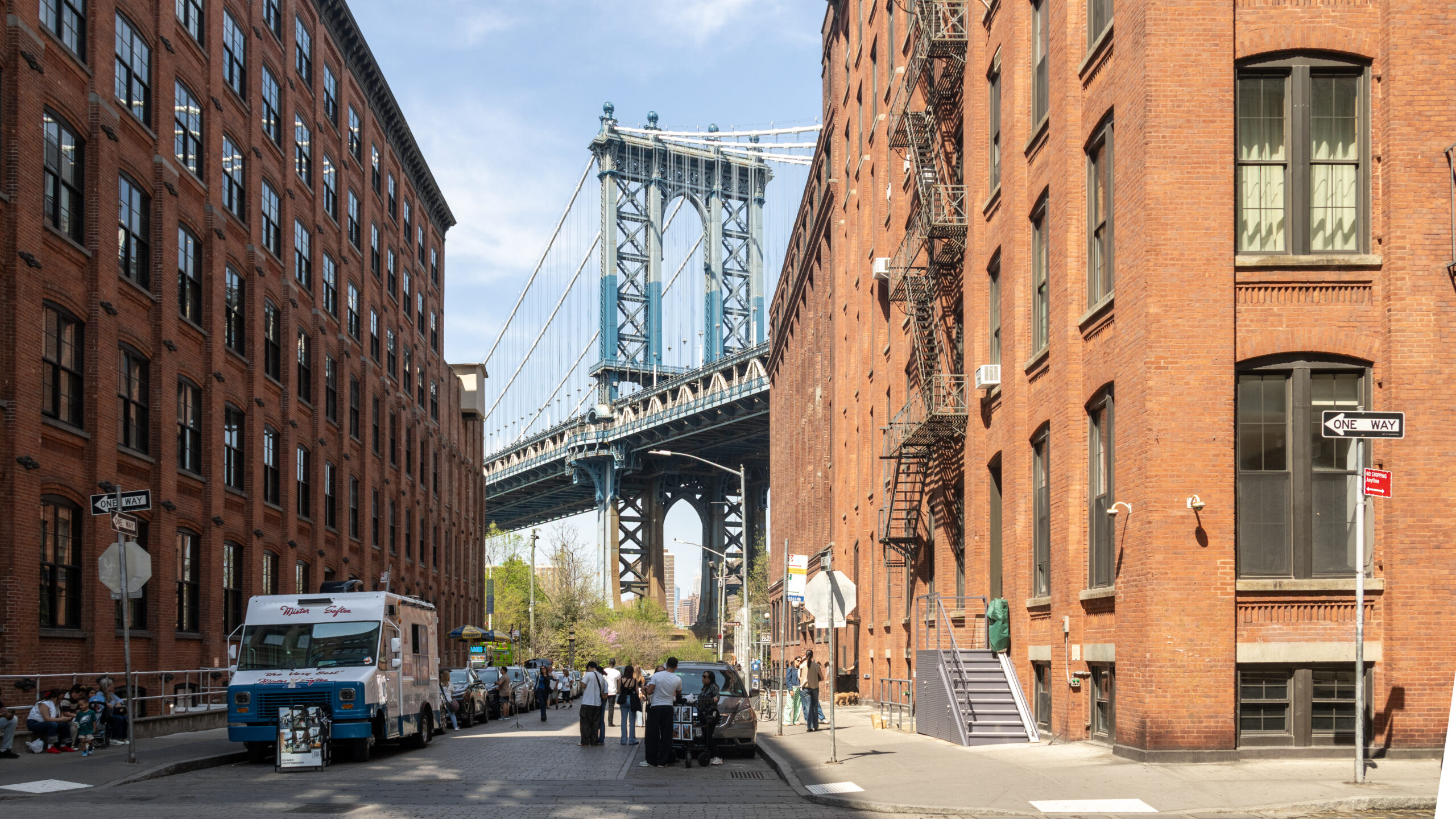 Brick buildings line a city street with the Manhattan Bridge visible between them.