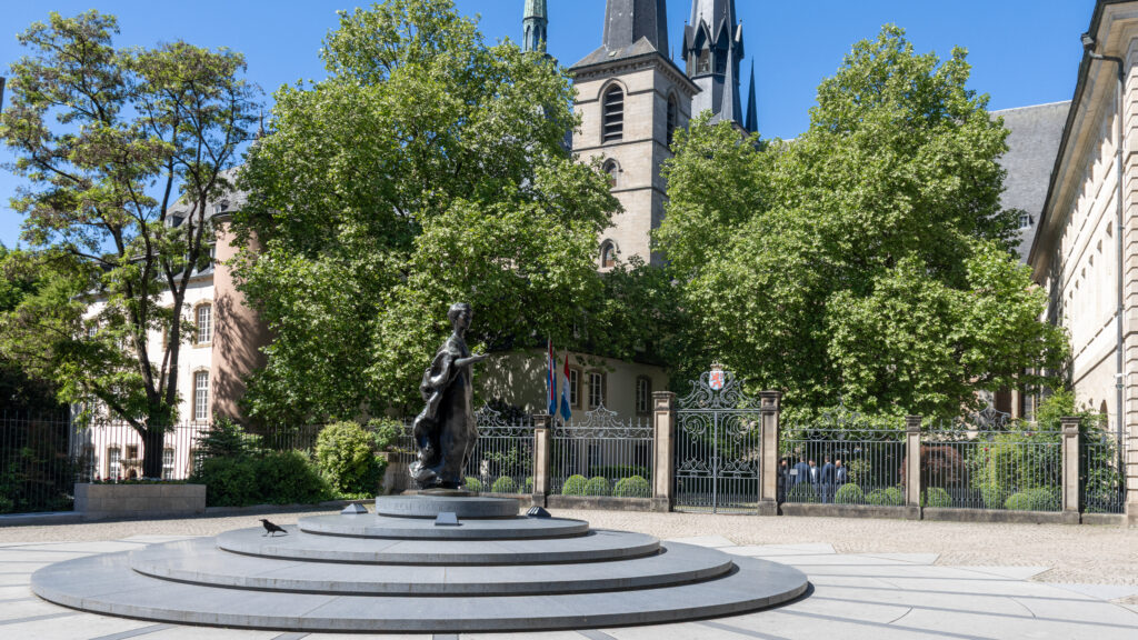 Place de Clairefontaine Luxembourg | Krista the Explorer Bronze statue in a circular, stepped plaza with a historic church.