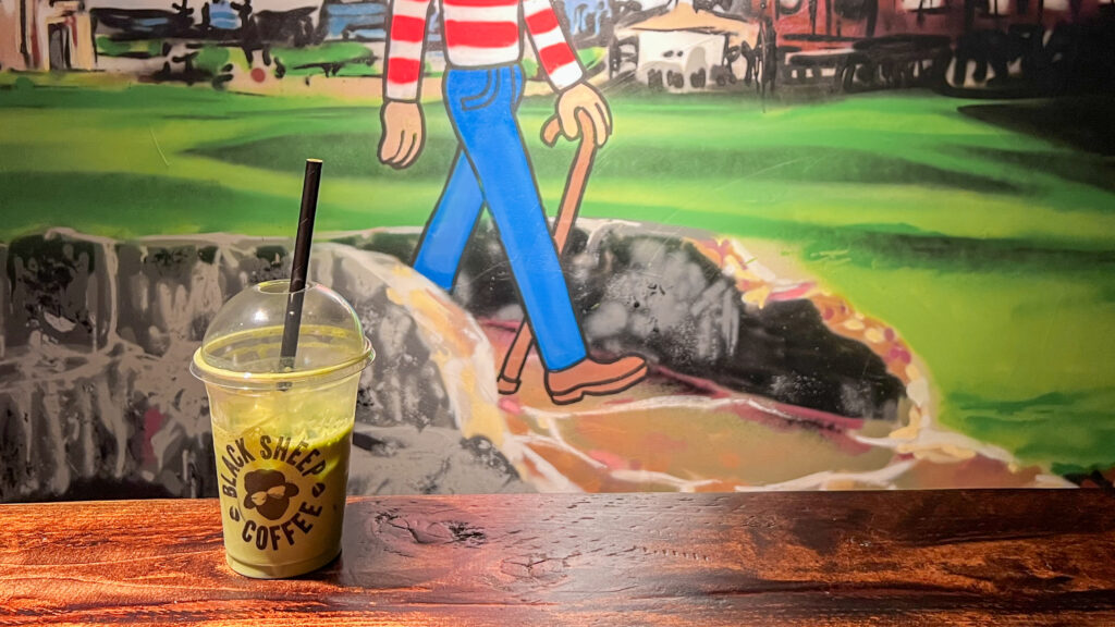 Iced matcha latte on wooden table with golf mural on wall.