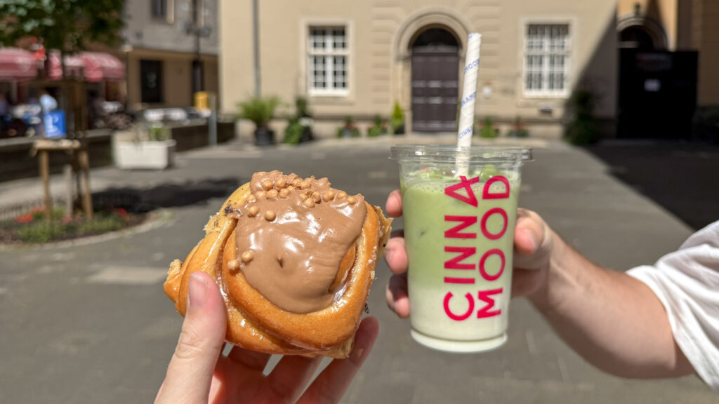 Cinnamood Luxembourg | Krista the Explorer Two hands holding cinnamon bun and iced matcha latte.