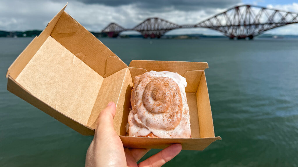 Cinnamon bun in a brown takeout box held up in front of bridge.