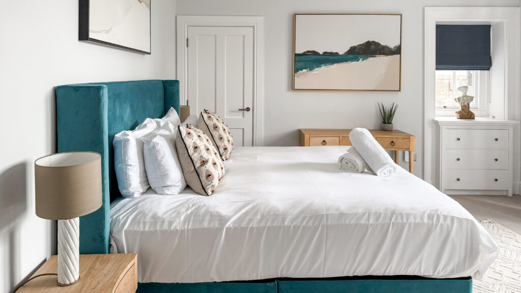 Teal upholstered bed with white bedding and patterned pillows in a bright, modern bedroom.