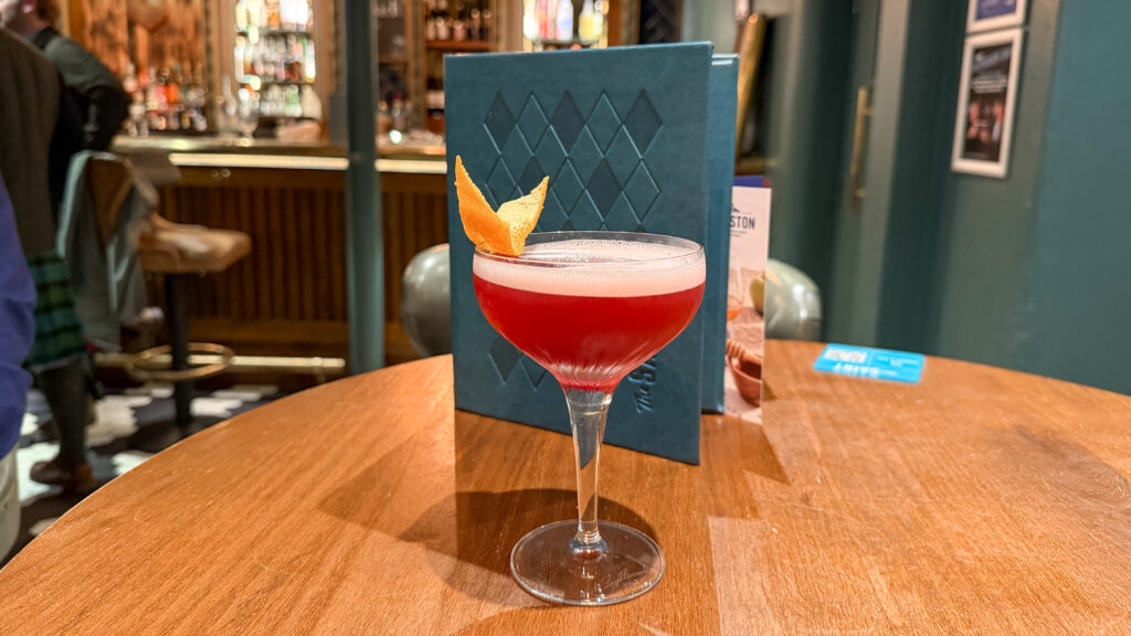 Red cocktail in a coupe glass with an orange peel garnish on a wooden bar table.