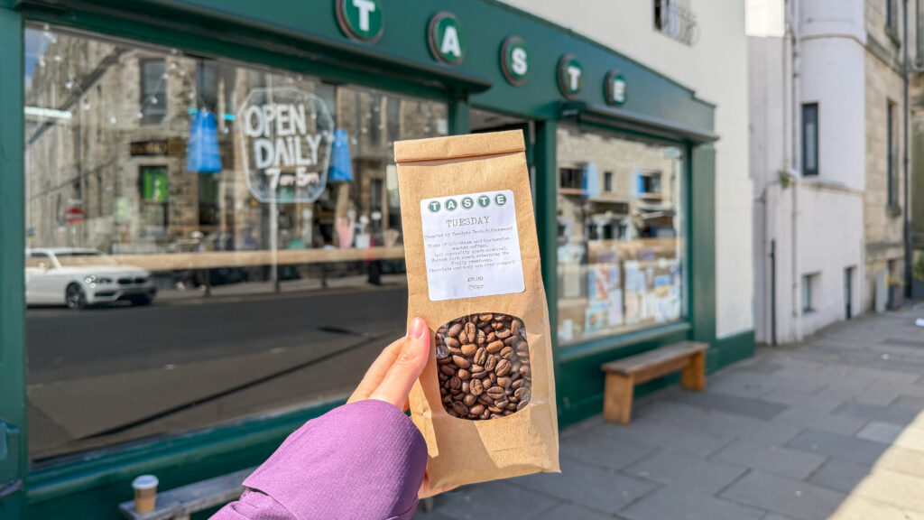 Person holding a brown paper bag of coffee beans in front of a cafe with a green exterior.