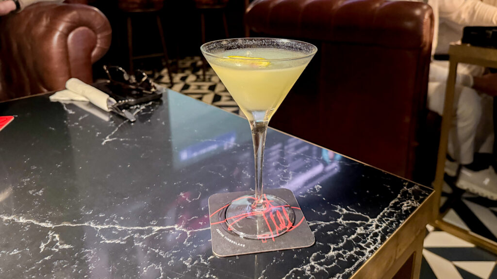 Martini glass with pale yellow cocktail and a lemon twist on a dark marble bar table.