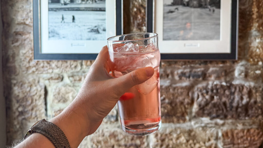 Glass of strawberry cider held up in front of stone wall.