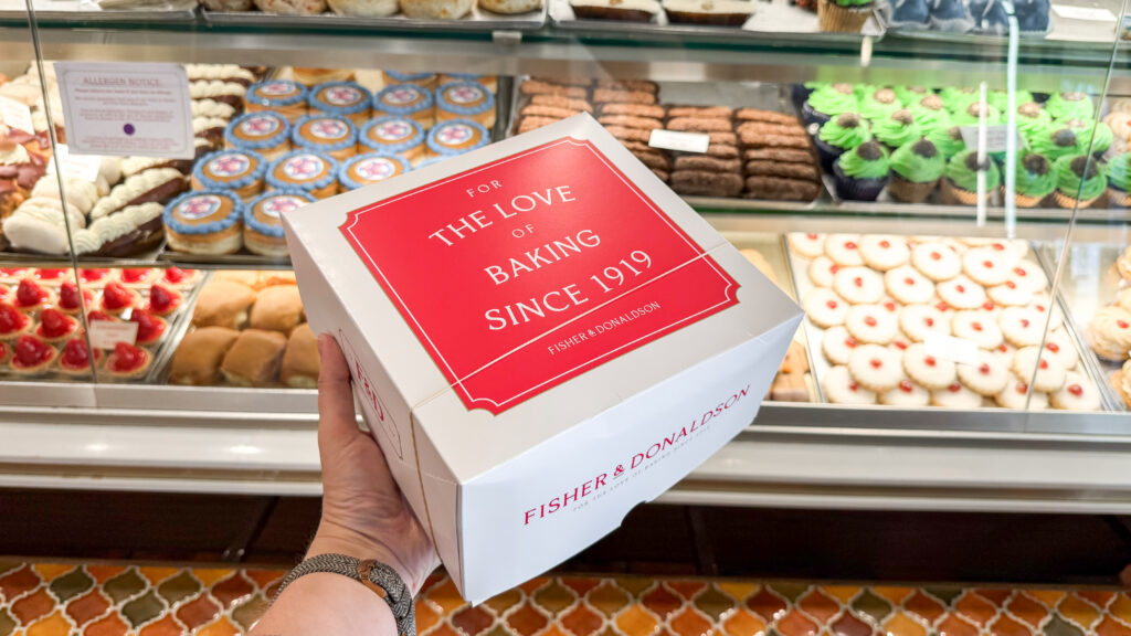 Person holding a white box with red label reading'For the Love of Baking Since 1919'.
