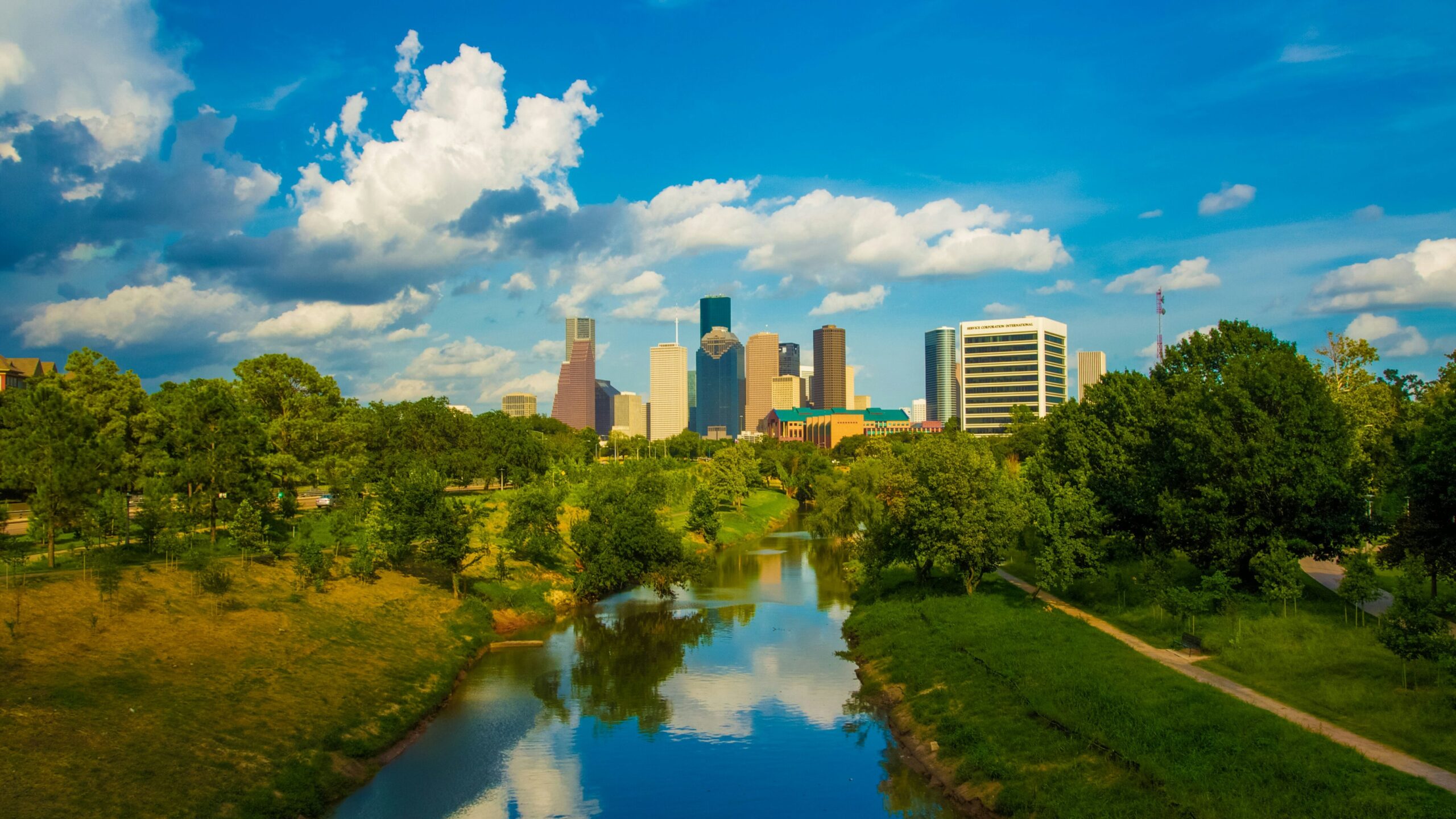 Downtown skyline across a calm river with green parkland in front.