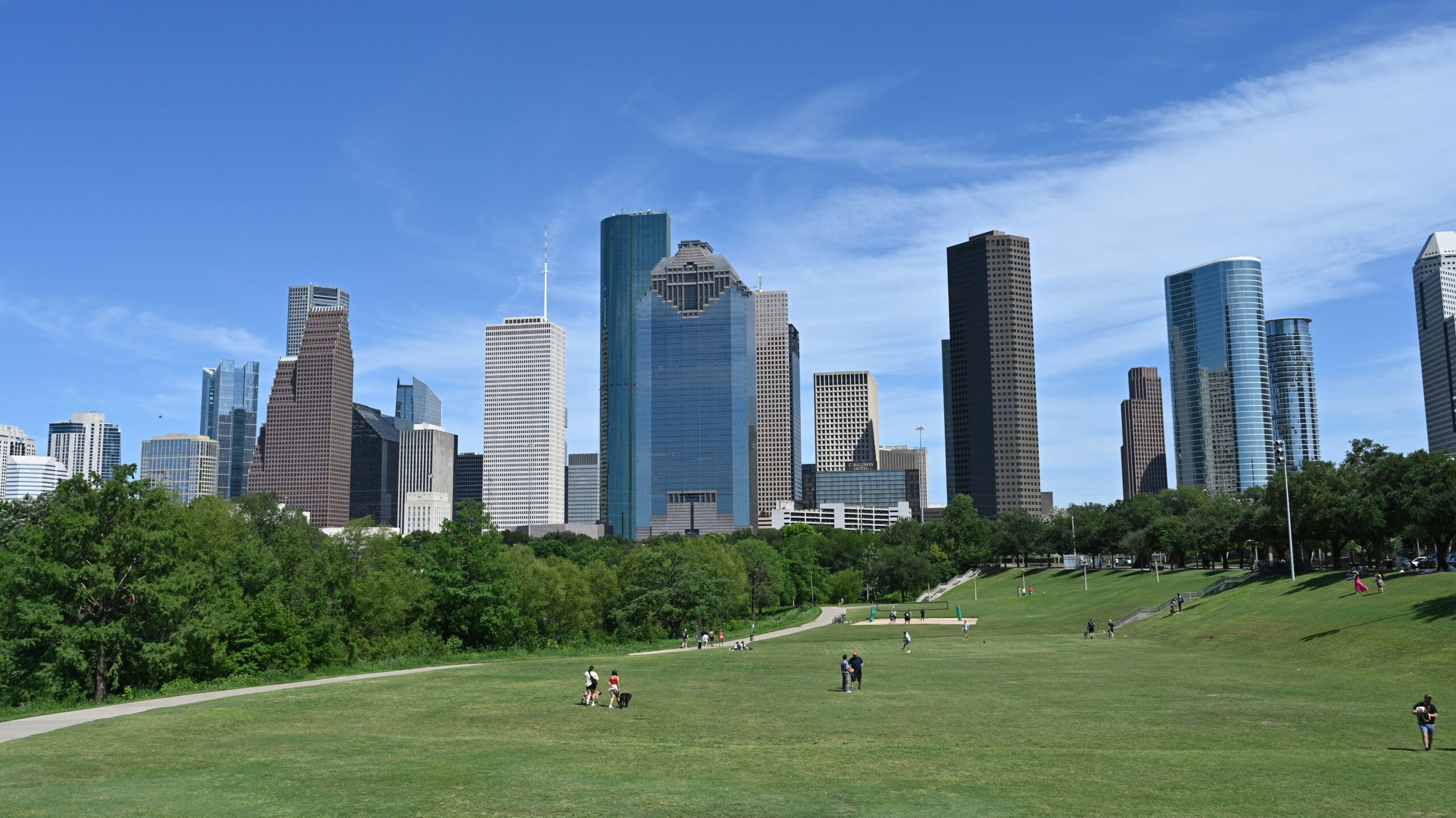 Skyscrapers rise behind a grassy park with people enjoying a sunny day.