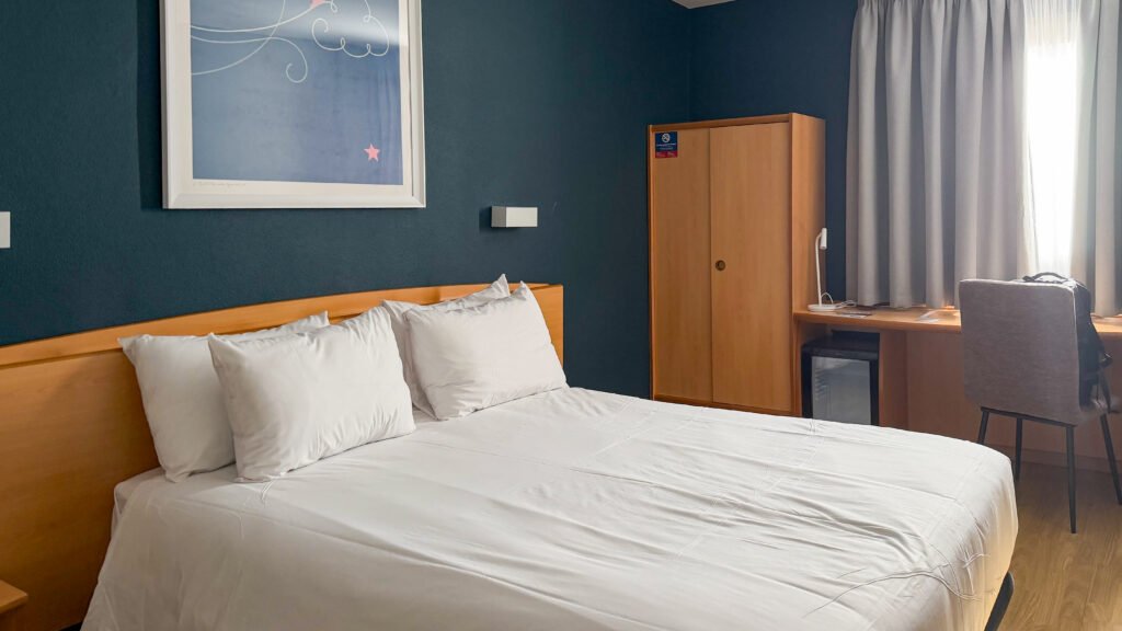 Travelodge Murcia | Krista the Explorer Hotel room with a large bed, white linens, dark blue accent wall, wooden wardrobe, and a desk beside a window.