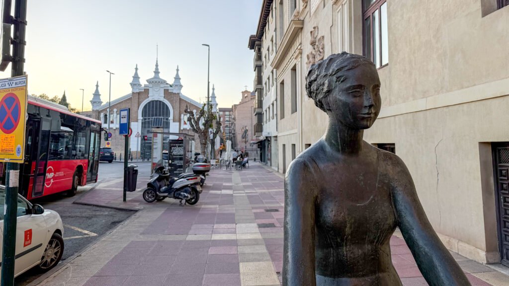 La Venus en Bicicleta Murcia | Krista the Explorer Bronze statue in the foreground on a city sidewalk with market behind.