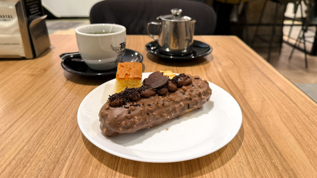 Chocolate eclair on plate with other pastries next to cup of tea.