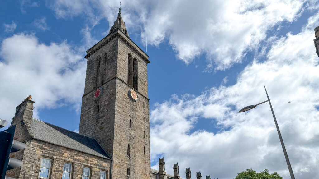 Stone church tower with a red clock face and a pointed spire against a bright blue sky.