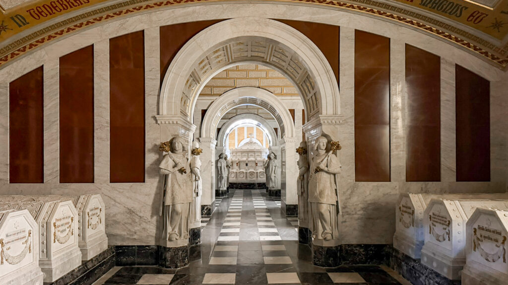 Marble mausoleum interior with repeating arches, statues in robes flanking a central corridor, and a checkered floor.