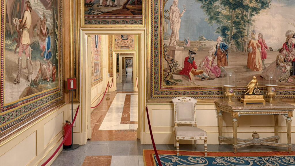Ornate palace hallway with large tapestries, gold-trimmed frames, and a decorative marble table.